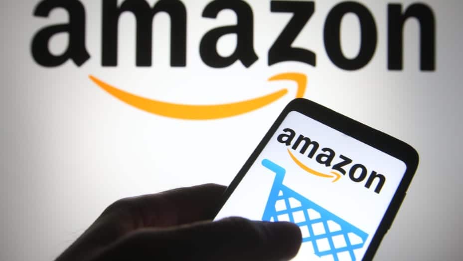 Logo d'Amazon en arrière-plan avec une main tenant un téléphone affichant le logo d'Amazon et une icône de panier d'achat sur l'écran.