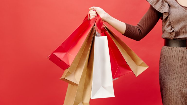 Une main tenant plusieurs sacs de shopping en papier rouge, blanc et kraft contre un fond rouge vif. L’image évoque l’univers du commerce, des achats et du retail.